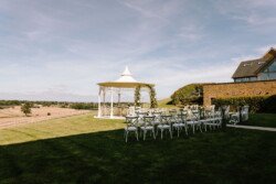 Outdoor wedding ceremony setup with white chairs facing a decorative gazebo under a clear sky at Croftview Weddings in Hinton Park. A field and building are in the background.