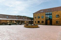 A courtyard with brick paving is flanked by a large two-story building and an adjacent single-story structure, both featuring tan brick and tile roofs. The central circular flower bed adds charm to this picturesque setting, perfect for Croftview weddings at Hinton Park.