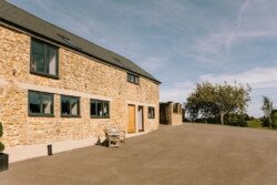 A stone farmhouse with a paved front yard, featuring several windows and a wooden front door. A single chair is placed near the entrance, and a tree and open fields are visible in the background. Perfect for Croftview weddings at Hinton Park, this location offers rustic charm in every corner.