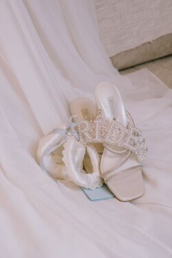 Pair of white bridal high-heeled sandals with crystal embellishments, one of which spells "BRIDE." Next to them lies a white lace garter with a blue ribbon on a white fabric backdrop, perfect for Croftview weddings.