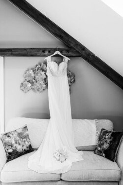 At Croftview weddings, a wedding dress hangs from a beam above a sofa, with a pair of shoes and a bridal bouquet placed on the cushion beneath it, creating an enchanting scene at Hinton Park.
