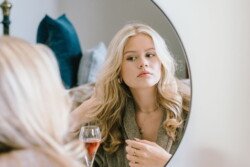 A woman with long blonde hair looks at herself in a round mirror, adjusting her hair with one hand. A glass of red wine is on the table beside her. Dressed in a grey robe and simple jewelry, she prepares for the Croftview weddings at Hinton Park.