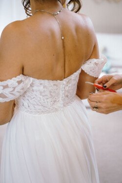 A bride in a white, off-the-shoulder, lace wedding dress is being assisted with buttoning the back of the dress at the enchanting Hinton Park.