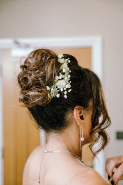 A woman with curly hair in an updo, adorned with small white flowers, wearing a necklace and earrings, faces away from the camera. Captured beautifully by Croftview Photos.