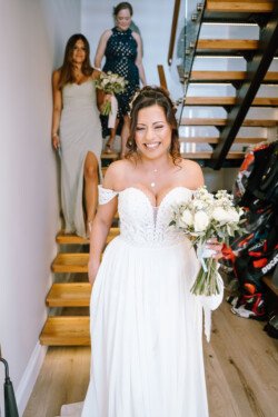 A bride in a white dress, holding a bouquet, walks down wooden stairs, followed by two women in dresses. The moment is beautifully captured by Croftview Photos.