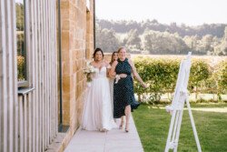A bride in a white dress and a woman in a blue polka dot dress walk along a path next to a building, smiling and carrying flowers. Captured perfectly by Croftview Photos.