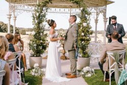 A bride and groom stand facing each other under a floral arch during an outdoor wedding ceremony, as guests seated on white chairs watch. Captured by Croftview Photos, a man in a suit stands to the side with a microphone.