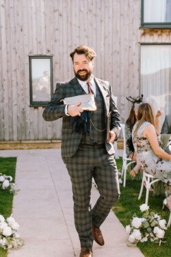 A man in a checked suit walks outdoors carrying a package, smiling. He is surrounded by people seated on white chairs with flowers on the ground. The background, beautifully captured by Croftview Photos, includes a wooden building.