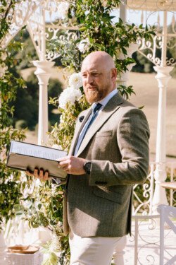 A man in a suit holds a binder and speaks in an outdoor setting decorated with flowers and greenery, captured beautifully by Croftview Photos.