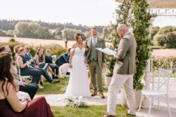 A wedding couple stands holding hands beside an officiant reading from a book outdoors. Guests are seated beside them, with trees and open fields visible in the background, all captured beautifully by Croftview Photos.
