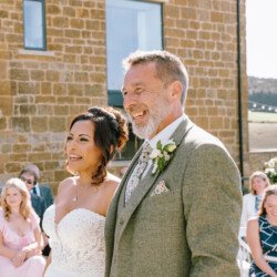 A bride in a white dress and a man in a gray suit stand outdoors, smiling. Guests are seated in the background near a brick building and countryside view, captured perfectly by Croftview Photos.