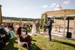A wedding ceremony is taking place outdoors on a sunny day. The bride and groom stand near an officiant reading from a book, while guests sit on white chairs under a gazebo. Croftview Photos captures these beautiful moments with stunning clarity and emotion.
