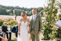 A bride and groom stand outside holding hands, smiling. The bride wears a white dress, and the groom wears a light grey suit. Captured perfectly by Croftview Photos, guests are seated in the background on a sunny day.