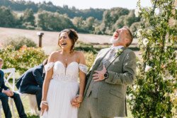 A bride and groom share a joyous laugh while standing outdoors during their wedding ceremony, captured beautifully by Croftview Photos, with guests seated behind them and a scenic landscape in the background.