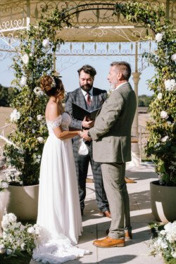 Bride and groom stand facing each other holding hands under a floral arch, with an officiant in the background during an outdoor wedding ceremony, beautifully captured by Croftview Photos.