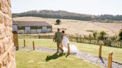 A couple dressed in wedding attire walks hand-in-hand down a path in a rural landscape with green fields, trees, and farm buildings in the background, beautifully captured by Croftview Photos.