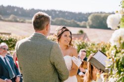 A bride and groom stand outside during their wedding ceremony, with a person holding a book in front of them and guests seated in the background on a sunny day. This beautiful moment was perfectly captured by Croftview Photos.