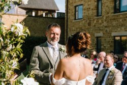 A man in a light gray suit and a woman in a white dress exchange vows during an outdoor wedding ceremony. Guests are seated in the background near a stone building, captured beautifully by Croftview Photos.