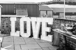 Large illuminated letters spelling "LOVE" are displayed against a stone wall outdoors, captured beautifully by Croftview Photos. Nearby, there is a table with a bowl of drinks and glasses. Industrial buildings and rolling hills can be seen in the background.
