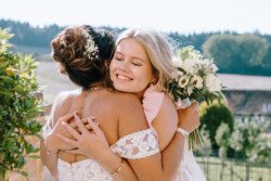 Two women embrace each other outdoors at a Croftview Weddings event at Hinton Park Estate, one holding a bouquet and the other wearing a strapless lace dress with floral hair accessories.