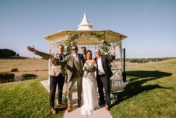 A bride and groom stand smiling under a gazebo at Croftview Weddings, surrounded by three men in suits, on a sunny day in the grassy field of Hinton Park Estate.