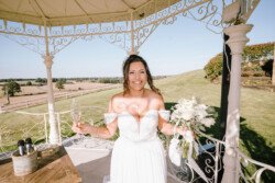 A woman in a white off-the-shoulder wedding dress stands under a gazebo at Croftview Weddings, holding a bouquet in one hand and a glass of champagne in the other, with the scenic landscape of Hinton Park Estate in the background.