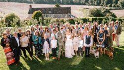A large group of people, dressed in formal and semi-formal attire, stand together outside on a lawn at Hinton Park Estate in front of a countryside house, with dogs also present. The scene at Croftview Weddings appears to be a celebration or gathering.