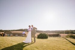 A couple in formal attire poses outdoors on a sunny day with clear skies, standing on a green lawn at Hinton Park Estate, with the charming barn and trees in the background. Perfect for capturing the essence of Croftview Weddings.
