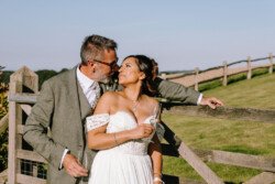 A couple dressed in wedding attire stand close together by a wooden fence at Hinton Park Estate, sharing an intimate moment outdoors under a clear sky. The man leans on the fence with his arm around the bride, capturing the elegance of Croftview Weddings.
