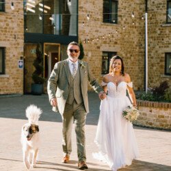 A bride and groom walk hand in hand outside, accompanied by a dog. The groom is in a grey suit, and the bride is in a white dress, holding a bouquet. They are smiling as they enjoy the beautiful setting of Croftview Weddings at Hinton Park Estate, framed by the charming rustic building.