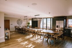A spacious dining area at Hinton Park Estate with sunlight streaming in, featuring a long wooden table with several chairs, a large wall clock, and modern lighting fixtures hanging from the ceiling—perfect for Croftview Weddings.