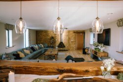 A spacious living room at Hinton Park Estate with a long grey sectional sofa, a wall-mounted TV, central modern fireplace, large windows, and wooden furniture. Pendant lights hang from the ceiling, perfect for relaxing after a day at Croftview Weddings.