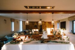 A table adorned with white linens holds a tiered white cake, cupcakes, flowers, and candles in a warmly lit room at Croftview Weddings, featuring modern decor and large windows at the beautiful Hinton Park Estate.