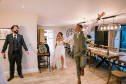 A man in a suit cheers while a bride in a white dress and another man in a suit smile at Croftview Weddings, set in the warmly lit Hinton Park Estate with its elegant long dining table.