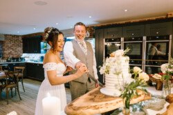 A couple in formal attire cuts a white layered cake in a modern kitchen with brick and dark cabinets at Croftview Weddings. White flowers and a wooden board decorate the scene, adding charm to their intimate celebration.