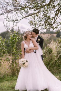 A couple in wedding attire stand outside under a tree at Orchardleigh Weddings. The groom embraces the bride from behind as she smiles, holding a bouquet.