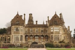 Historic stone mansion with multiple chimneys, ornate windows, and a grand staircase leading to the entrance, surrounded by a manicured lawn perfect for Orchardleigh House Weddings.