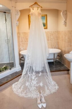 An elegant white wedding dress with detailed lace and a long veil displayed in a bathroom at Orchardleigh House, with shoes placed in front.