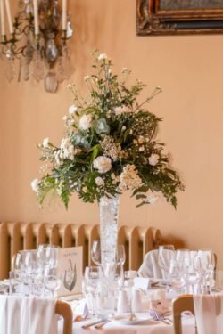 Tall floral centerpiece with white blooms and green foliage in a clear vase on a dining table set for an Orchardleigh House Wedding, with elegant place settings and soft lighting.