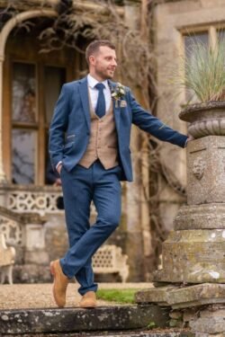 Man in a blue suit and vest with a boutonniere, leaning on a stone pillar outside Orchardleigh House, looking away happily.