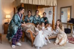 Women in robes and pajamas with young girls in white dresses gather in a bedroom at Orchardleigh House, smiling and interacting around an antique chair.