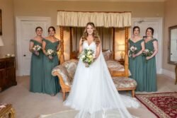 A bride in a white gown stands smiling with three bridesmaids in teal dresses, holding bouquets in an elegantly furnished room at Orchardleigh House.