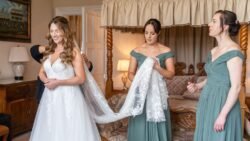 A bride in a white dress smiles as two bridesmaids in green dresses adjust her veil in an elegant bedroom at Orchardleigh House.