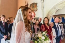 Bride smiling as she walks down the aisle at Orchardleigh House, holding a bouquet, with guests watching in a church adorned with paintings.