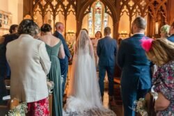 A bride in a white gown and veil walks down the aisle of Orchardleigh House, observed by guests in formal attire.