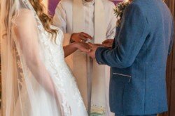 Bride and groom exchanging rings during an Orchardleigh House wedding ceremony, officiated by a priest in a white robe.