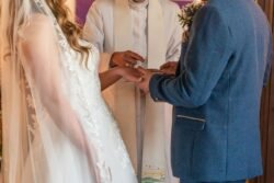 Bride and groom exchanging rings during an Orchardleigh House wedding ceremony, officiated by a priest in a white robe.