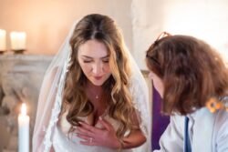 A bride in a white dress and veil listens intently to another woman indoors at Orchardleigh House, with candles illuminating the scene.