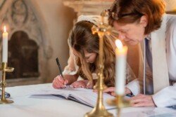 At an Orchardleigh House Wedding, a young woman in a veil signs a document in a church beside an older woman, with lit candles on either side.
