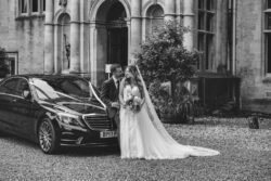 A newlywed couple standing beside a luxury car in front of Orchardleigh House, sharing a romantic moment. Black and white photo.